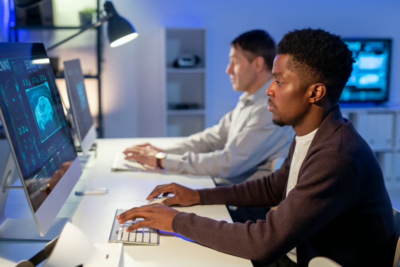 Black technology professional working at a computer with digital data visualizations while a colleague works nearby.